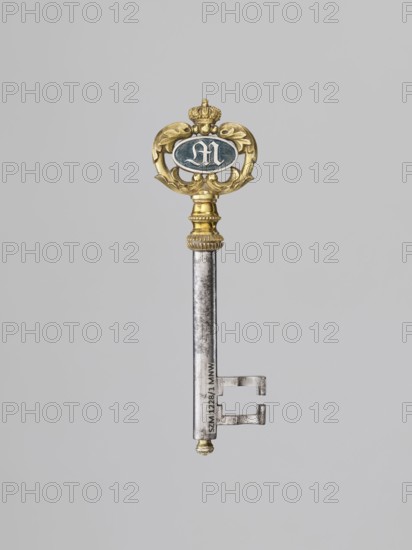 Souvenir key, steel, with a full shank and a simple feather in the form of two square brackets. Gilded bronze knot and handle: oval handle composed of two U-shaped sections decorated with leaves, fastened with a ducal mitre. Inside, an oval plaque with a Gothic monogram M and blue enamel on one side, and a cast inscription on the other: VON / DER STADT / DARMSTADT / DEN 26 DECEMB: / 1833. Given together to Duchess Mathilde Caroline of Bavaria 1813–1862 and her husband, the future ruler of Hesse, Ludwig III, on the occasion of their wedding by the city of Darmstadt, 1833. Germany; Darmstadt. Varia. Height 8.9 cm, width 2.6 cm., A single ornate key is shown against a plain light gray background, featuring a decorative top with gold tones and a small crown element, an oval medallion with a dark bluish area, and an elongated silvery metal shaft with a few notches at the biting end; the visible colors include gold, silver, dark blue, gray, and a hint of darker metallic patina.
