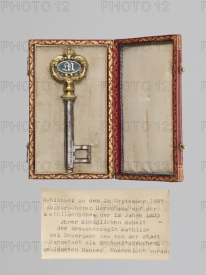Souvenir key, steel with full shank and a simple feather shaped like two square brackets; gilded bronze knot and handle: oval handle of two U-shaped leaf-decorated sections fastened with a ducal mitre. Inside, an oval plaque with a Gothic monogram M and blue enamel on one side, and a cast inscription on the other: VON / DER STADT / DARMSTADT / DEN 26 DECEMB: / 1833. Given to Duchess Mathilde Caroline of Bavaria 1813–1862 and her husband Ludwig III on the occasion of their wedding, 1833, Darmstadt, Germany; height 8.9 cm, width 2.6 cm., A decorative metal key rests in an open rectangular box lined with light fabric; the box exterior rim is red with gold ornamental trim and the interior edges have a blue twisted cord, while the fabric interior is a pale beige. The key has a long metal shaft that appears silver-gray and a more ornate bow with gold-toned elements and a darker oval center featuring a pale design. The box is shown against a plain light gray background. Below the box there is...