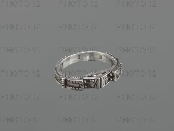 Bracelet, maker, scrap metal; 19th century, China/Mongolia; dragon with pearl motif; Mongolian/Chinese decorative style; body/hand ornament, fashion accessory; hundred antiquities baigu ornament; materials: jewelry; dimensions: height 8.0 cm, width 9.0 cm, weight 281 g., A silver-colored ornate ring sits centered against a solid gray background, showing detailed raised patterns and engraved motifs along its outer band; the ring has a slightly weathered metallic sheen with darker patina in recessed areas and smoother, lighter highlights on raised surfaces, and the design includes repeating scroll-like decorations and textured panels that wrap around the circular shape.