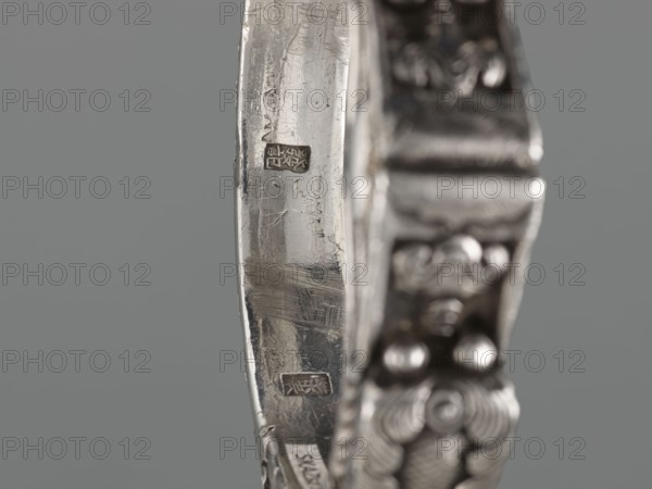 Bracelet, scrap metal, purchased; Chinese and Mongolian art influences; body/hand ornament, fashion accessory; motif: dragon with pearl; hundred antiquities baigu ornament; culture: Chinese; 19th century, origin: China, and Mongolia ; jewelry; height 8.0 cm, width 9.0 cm, weight 281 g, A close-up of a metallic object with a curved band and ornate edge, showing worn and scratched surfaces in silver and gray tones, with a blurred decorative side featuring rounded elements and textured patterns in darker silver; the background is a flat muted gray.