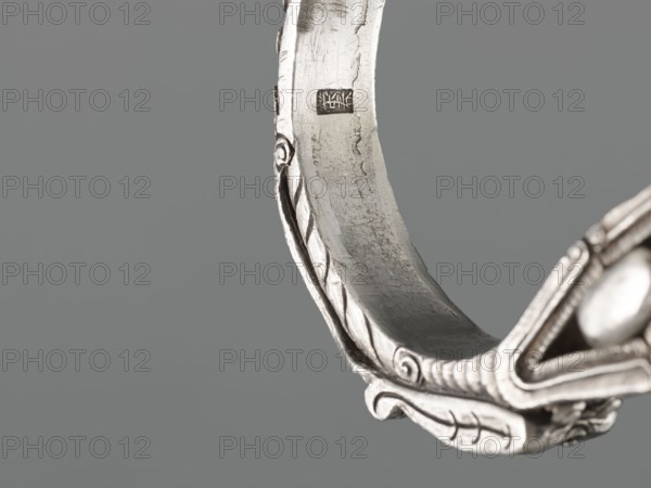 Bracelet, scrap - purchase, purchase provenance; Chinese and Mongolian art; body/hand ornament jewelry, fashion accessory; dragon with pearl motif, hundred antiquities baigu ornament; 19th century, China, Mongolia ; height 8.0 cm, width 9.0 cm, weight 281 g, The image shows a close-up of a curved metallic object with ornamental raised patterns and scroll-like details, presented against a smooth grey background; the metal appears silver with areas of darker tarnish and subtle reflections, and there is a rounded bead-like element visible at one edge.