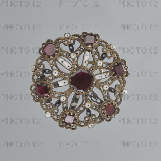 Earring, mid-19th century, Italy, jewelry, diameter 2.6 cm, A round ornate brooch with an open filigree metal framework in a brass or gold tone, featuring a central deep red faceted stone surrounded by a group of several smaller deep red faceted stones set around the perimeter, interspersed with elongated white enamel elements that have small dark speckles and a few small round white enamel dots, and additional small dark teardrop-shaped accents; the overall color palette includes gold or brass, deep red, white, black or dark gray, and the background is a pale gray.