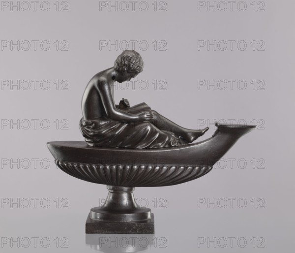 Decorative cast-iron oil lamp, 19th century, Poland; attributed to Szewczykowski, Roman 1849–1901. Square plinth supporting an oval, boat-shaped container with a slightly raised end and a flat hinged lid. A young man sits on the lamp, leaning over an open book. Modeled on a French design possibly Thomire and known in several copies; likely cast in Poland casting quality lower than some other known examples. Unlike other examples, there is no hole in the cover closing the opening in the body. These objects were mostly decorative e.g., for a fireplace and not suitable for actual lighting due to weak capillarity of oil in deep fuel tanks. Materials: cast iron. Function: decorative lighting object. Dimensions: height 28.5 cm; width 11 cm; depth 33.5 cm., A dark bronze sculpture of a seated figure in a draped garment reclining inside a boat-like basin mounted on a fluted pedestal, the figure shown in profile with head bowed and arms resting on knees, rendered with textured hair and smooth m...