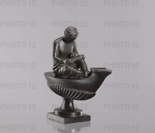 Decorative cast iron oil lamp, cast by Szewczykowski, Roman 1849–1901. 19th century, Poland. Square plinth supporting an oval boat-shaped container with a slightly raised end and a flat hinged lid. A young man sits on the lamp, leaning over an open book. Modeled on a French design possibly Thomire known in several copies; similar examples made at various workshops and foundries including glassworks and royal Prussian foundries though casting quality varies — this example’s casting is of lower quality and likely cast in Poland. Unlike other examples, there is no hole in the cover closing the opening in the body; these objects were decorative e.g., for a fireplace and not suitable for lighting. Materials: decorative cast iron. Function: lighting decorative. Dimensions: height 28.5 cm; width 11 cm; depth 33.5 cm., A dark bronze-colored sculpture of a seated figure reading sits atop a boat-shaped basin with a fluted lower half, all mounted on a short pedestal and square base; the figure ha...