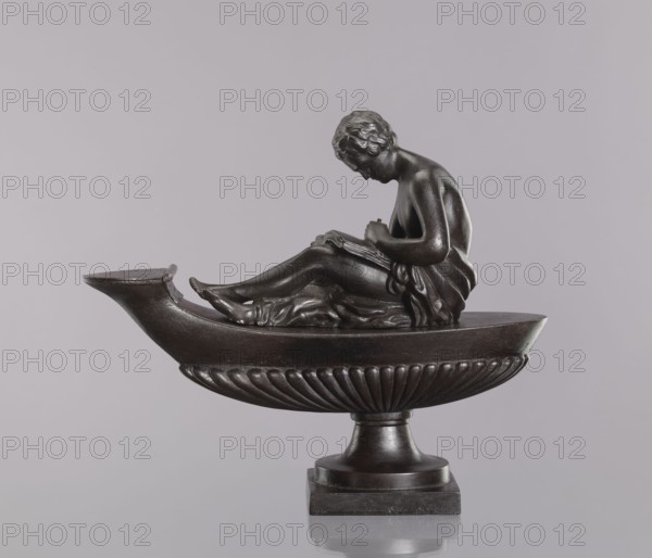 Decorative cast-iron oil lamp, attributed to Szewczykowski, Roman 1849–1901. Square plinth supporting an oval boat-shaped container with a slightly raised end and a flat hinged lid; a young man sits on the lamp, leaning over an open book. Modeled on a French design perhaps Thomire and known in several copies; likely 19th century, probably cast in Poland possible workshops: Aleksandra glassworks in Bialogon, Drews in Warsaw, or Suchedniów glassworks; less likely the Minter family or Prussian royal foundries. Casting quality is relatively low compared with other known examples; unlike other examples, there is no hole in the cover closing the opening in the body. These lamps were decorative elements e.g., for a fireplace and were not suitable for actual lighting due to weak capillarity of oil in such deep fuel tanks. Materials: cast iron. Function: lighting/ornamental. Dimensions: height 28.5 cm, width 11 cm, depth 33.5 cm. Location: 19th-century Poland, possibly Warsaw., A dark bronze-co...
