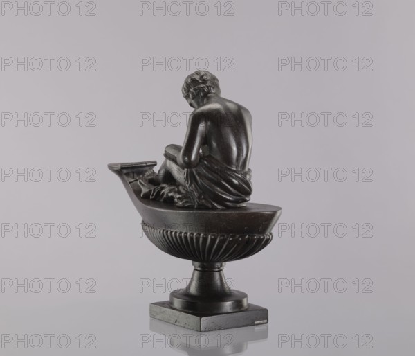 Decorative cast iron oil lamp, 19th century, Poland; square plinth with an oval boat-shaped container, slightly raised end and flat hinged lid. A young man sits on the lamp, leaning over an open book. Modeled on a French design possibly Thomire, known in several copies; likely decorative rather than functional deep fuel tank unsuitable for lighting; no hole in the cover to vent the body. Casting quality lower than some identical examples; possibly cast in Poland Aleksandra glassworks in Bialogon, Drews in Warsaw, or Suchedniów glassworks have been suggested, with replicas also produced at Prussian foundries Gliwice, Berlin. Height 28.5 cm, width 11 cm, depth 33.5 cm., A dark bronze-colored figurine of a seated person viewed from behind, positioned on a boat-shaped bowl with fluted sides atop a pedestal, all resting on a square base; the figure wears draped cloth around the lower body and has short wavy hair, with visible smooth contours and reflective highlights. The background is a pa...