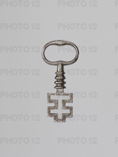 Key, Szewczykowski, Roman 1849–1901 — Key for a ratchet lock with a feather at the end of the shaft forming an outline of a cross on a horizontal beam. The shaft has profiles and an oval handle. This type of lock was already in use in the Roman period. The keyhole is in the shape of an inverted T; opening is achieved by vertical movement of the key, not rotation. In France it is called à la cordelière or à la capucine due to depictions of such a key hanging at the end of a cord used to tie Capuchin habits; this type of lock was often used in monastery cell doors. The bolt mechanism is similar to locks used in rural buildings into the 20th century, functioning as a handle lock opened by a lever passing through the door and ending on the other side with a flat plate pressed with the thumb. 18th century; locksmith. Dimensions: height 6.6 cm, width 2 cm, depth 0.9 cm., A single metal key with a rounded oval handle and a short grooved stem, showing a textured, slightly worn surface in shade...