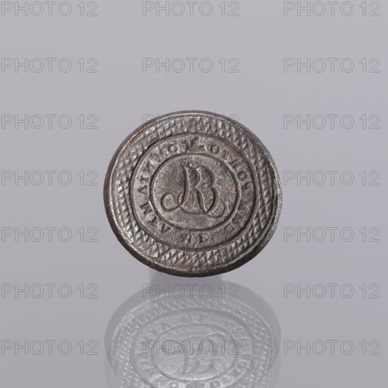 Seal stamp, engraving. Oval, almost circular; central field with engraved monogram ER R mirror-reversed. Rim surrounding central field embossed with punches reading IMMPORTO image of a bird in flight OMNIA MEA MF. Outer rim engraved with a diagonal grid. Stamp in the form of a plate with a cylindrical sleeve on the reverse for mounting a wooden handle. Early 19th century, Europe. Height 3 cm, width 3.2 cm, depth 2.4 cm., A round metallic object with a textured rim and a circular raised design in the center sits against a pale gray background; the metal appears dark silver and brownish with a slightly worn surface, and a soft reflection of the object is visible below it showing similar dark silver and gray tones.