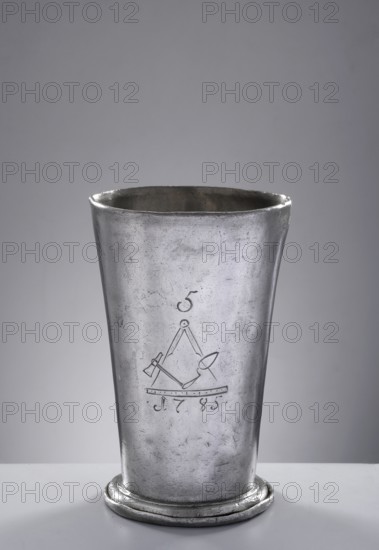 Bricklayers' guild mug. Tin mug of conical shape, set on a profiled convex base with a turned-down rim, with a smooth casing; in the center masonry emblems: compass, trowel, hammer, and square with the number 5 above and the date 1785 below. Circa 1785. Poland, Royal Prussia, Chojnice . Guild memorabilia; vessel. Diameter 9.8 cm; height 15.5 cm., A tall metallic cup with a slightly flared rim and a rounded base sits on a flat surface; the cup is silver-gray in color with a worn, scratched texture and subtle darker gray areas, and the background is a smooth, light gray gradient.
