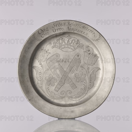 Plate by Otto Friedrich Groth fl. 1792–1822. Flat pewter plate with a smooth mirror, semicircular notch, and narrow upturned collar. In the mirror is an engraved representation of unidentified crossed guild tools with the date 1812 possibly meat cleavers, surrounded by a stylized plant twig with symmetrically arranged flowers, including tulips on leafy stems, beneath a crown. On the collar is an engraved inscription surrounded by leafy branches: CARL. STOSZ. BEYSITZ-MEISTER. / CARL OTTO ALTGESTELLE., 1812. Brzeg, Silesia. Plate; height 2.2 cm, width 22.2 cm., A round metal plate with a raised rim, shown against a pale background, featuring an engraved central design of two crossed objects surrounded by floral motifs and decorative scrollwork, with a crown-like element above and stylized flowers and leaves flanking the central motif; the plate has a satin metallic surface with areas of darker and lighter gray.