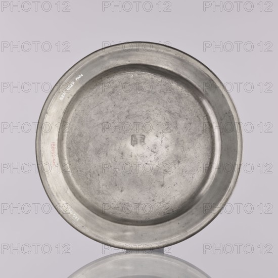 Plate by Otto Friedrich Groth fl. 1792–1822. Flat pewter plate with a smooth mirror, semicircular notch, and narrow upturned collar. In the mirror is an engraved representation of unidentified crossed guild tools dated 1812 possibly meat cleavers, surrounded by a stylized plant twig with symmetrically arranged flowers including tulips on leafy stems, beneath a crown. On the collar is an engraved inscription surrounded by leafy branches: CARL. STOSZ. BEYSITZ-MEISTER. / CARL OTTO ALTGESTELLE., 1812, Brzeg, Silesia. Plate: height 2.2 cm, width 22.2 cm., A round shallow metal plate with a raised rim shown from above, its surface marked by scratches and areas of uneven texture, with a small shallow indentation near the center; the plate appears in shades of gray and silver, set against a pale muted lavender-gray background, and a soft reflected oval of the plate is visible beneath it in similar gray tones.