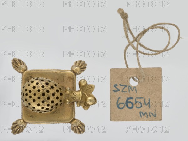 Toothpick holder, hedgehogs, animals, tassels; in the form of a hedgehog on a pad with tassels on the corners; 2nd quarter of the 19th century; Poland, possibly Warsaw; varia; height 3 cm, width 4.3 cm, depth 4.7 cm, A small metallic object with a golden finish sits on the left, showing a square base with decorative rounded corner feet and a perforated dome attached to one side topped by a small sculpted element; to the right a rough rectangular kraft-colored tag hangs from a loop of coarse light-brown twine, the tag showing some blue markings; background is a pale light-gray.