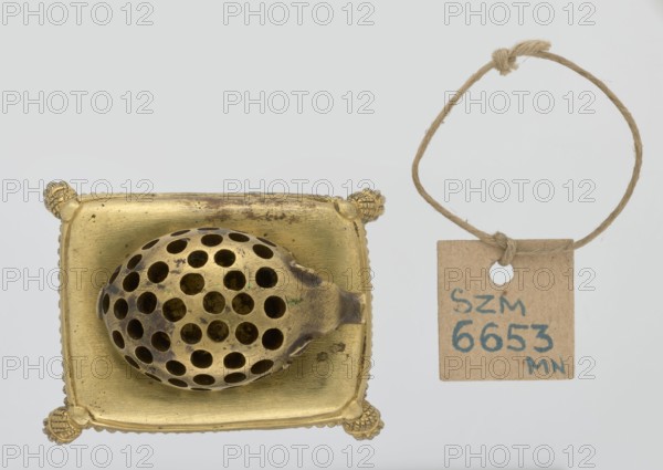 Toothpick holder, hedgehogs, animals, tassels. Bronze toothpick holder in the shape of a pig resting on a rectangular base bordered with a fringe. 1820s–1840s, Poland . Varia. Height 3.5 cm; width 6.3 cm; depth 4.7 cm., A metallic object with a golden color sits on a square golden base with decorative rounded corners; on the base rests an oval perforated piece with many round holes and some darker areas of patina. To the right, a small square tag made of brown paper hangs from a loop of coarse beige twine; the twine forms a loop with a knot at the top. The background is a very light gray.