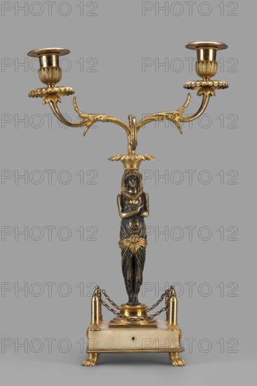 Two-candle candelabra depicting Egyptian women, early 19th century, France, unknown locality; lighting; height 35 cm, width 22.5 cm, depth 11.3 cm, A decorative two-armed candlestick featuring a central sculpted female figure with crossed arms, standing on a rectangular base; the piece combines dark metallic and bright gold tones with a light stone-colored base, a group of ornate gold arms extending outward with leaf-like motifs and two candle holders at the ends, a gold floral element at the figure’s waist, small chained railings connecting four gold posts around the base, and gold paw-shaped feet supporting the stone-colored platform.