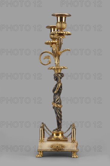 Two-candle candelabra depicting Egyptian women, early 19th century, France, unknown locality; function: lighting; height 35 cm, width 22.5 cm, depth 11.3 cm., The image shows a tall decorative candlestick with a vertically oriented figure of a draped female form serving as the central column, finished in dark bronze tones with gold highlights; the upper section branches into ornate floral and leaf motifs with a few petal-like bobeches and candle cups rendered in bright gold; the base is a square platform in a pale creamy color with gold decorative mounts, small gold feet shaped like paws, and short gold posts connected by delicate gold chains; the overall color palette includes gold, dark bronze, pale cream, and a gray background.