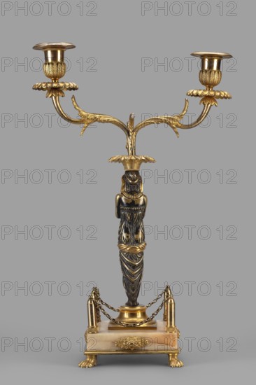Two-candle candelabra, Egyptian women, early 19th century, France, unknown locality, lighting, height 35 cm, width 22.5 cm, depth 11.3 cm, A gilded two-armed candlestick with a central vertical figure-like column standing on a rectangular base; the candlestick arms curve outward and upward with leaf-like decorative elements and floral drip pans beneath each socket, all in a gold tone, while the central column has a darker, patinated surface with carved drapery-like detailing and touches of gold accents; the rectangular base appears to be a light beige or cream stone with gold mounts at the corners, small chained railings in gold between short posts, and gold ornamental elements on the sides, all supported by four gold paw-shaped feet, set against a plain gray background.