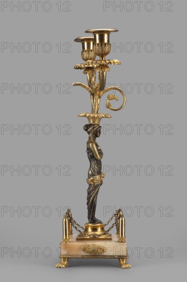 Two-candle candelabra, depicting Egyptian women, early 19th century, France, lighting, height 35 cm, width 22.5 cm, depth 11.3 cm, A tall decorative object stands against a plain grey background, featuring metallic gold and dark patinated bronze tones, with a base that shows creamy beige and light brown hues; the piece rises from a square plinth with clawed feet and a decorative gold emblem, topped by short posts linked with gold chains. The central shaft is formed by a sculpted female figure in dark bronze with gold accents in the drapery and around the waist, supporting above her head a flared gold element from which stylized leafy gold branches extend. At the top, these branches hold a group of ornate gold candle holders with textured and fluted surfaces and scalloped rims, and one curved gold scroll detail extends outward from the middle section. The overall palette includes gold, dark bronze, beige, light brown, and grey.