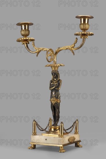 Two-candle candelabra depicting Egyptian women, early 19th century, France, unknown locality; for lighting; height 35 cm, width 22.5 cm, depth 11.3 cm., The image shows an ornate two-armed candelabrum with a central figure serving as the stem, mounted on a rectangular base; the piece features metallic gold surfaces and darker bronze tones, with a pale off-white base. The two upward-curving arms end in scalloped bobeches and cylindrical candle cups, all in gold, and the arms have leaf-like decorative elements in gold. The central stem is formed by a stylized female figure in a darker bronze color with gold accents at the waist and head, standing beneath a flared gold capital that supports the arms. The rectangular base is a pale off-white stone with gold mounts at the corners and small decorative gold feet shaped like paws, and a group of short gold posts linked by short dark metal chains surrounds the figure on the base. The overall palette includes gold, dark bronze, and pale off-whit...