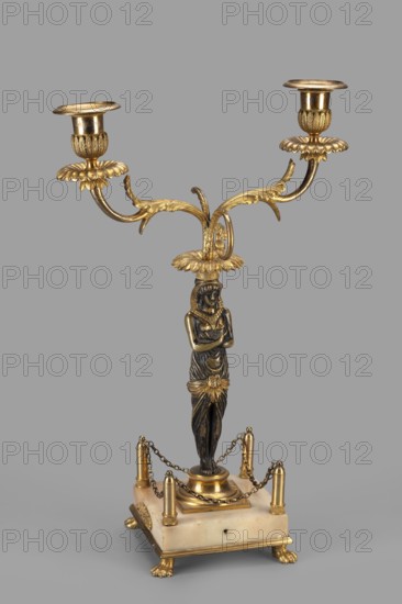 Two-candle candelabra, depicting Egyptian women, early 19th century, possibly France, lighting; height 35 cm, width 22.5 cm, depth 11.3 cm, A decorative candlestick with a metallic finish in gold and dark bronze tones stands on a square pale base with small claw feet; a sculpted female figure in dark bronze tones forms the central column, wearing draped garments and gold accents, and above her head a gold leafy element branches into two ornate curving arms each ending in a gold candle holder with petal-like bobeches; gold chains drape between small gold posts at the corners of the pale base, and the background is a uniform grey.