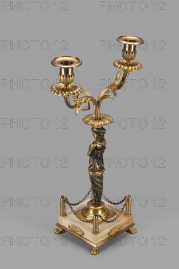 Two-candle candelabra, depicting Egyptian women, early 19th century, France, lighting, height 35 cm, width 22.5 cm, depth 11.3 cm, A decorative candlestick with a metallic finish in gold and dark bronze tones stands on a square base of pale stone, the base accented with gold ornamental plaques at its sides and small paw-shaped feet. A group of short gold posts at the base corners are linked by delicate gold chains. Rising from the center is a sculpted figure wrapped in draped clothing rendered in dark bronze with gold highlights, supporting a flared gold floral platform from which curved gold stems extend into a branching floral design topped by two gold candle holders, each with petal-like collars; the visible colors include gold, dark bronze, pale stone, and subtle variations of gray in the background.