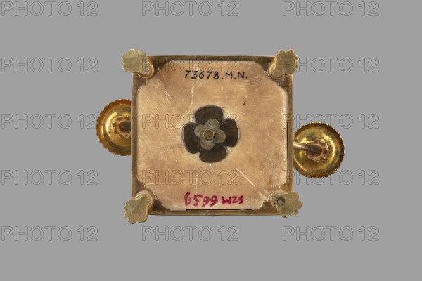 Two-candle candelabra depicting Egyptian women, early 19th century, possibly France, lighting, height 35 cm, width 22.5 cm, depth 11.3 cm, A square metallic object with rounded corner cutouts and a central layered flower-shaped detail, mounted on a gray background; the metal shows a mix of gold and brass tones with darker gray in the central flower piece and some lighter worn areas; decorative scalloped circular knobs extend from two opposite sides in gold tones; a few clover-shaped corner mounts in a dull gold color sit at each corner; the surface shows scratches and slight discoloration with visible highlights and shadows.