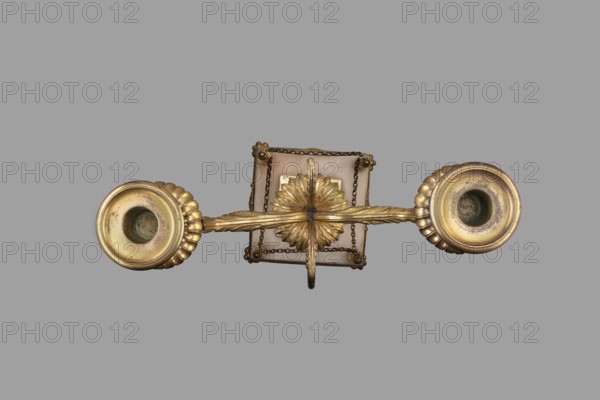 Two-candle candelabra depicting Egyptian women, early 19th century, France, lighting, height 35 cm, width 22.5 cm, depth 11.3 cm, A decorative metallic object with a central rectangular plate and a radial floral medallion, rendered in a gold tone with visible patina and darker tarnish; a textured horizontal bar with leaf-like grooves extends outward to two circular cup-like ends that have scalloped outer rims and darker inner cavities; a short hanging loop projects downward from the center and small chain links are draped around the rectangular plate; the background is a uniform gray.