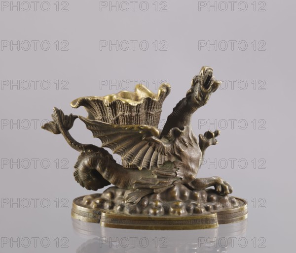 Spirit lamp, In the form of a two-winged dragon on a four-leaf oval base. Between the wings is a container; in the mouth is a hole for the wick, and on the side is a hole for pouring spirit. According to information in Szewczykowski’s catalogue, this is an oil lamp and a very old one. Due to the relatively large distance from the bottom of the container to the mouth, the lamp-burner cannot be fired with oil, which has poor capillarity. Most likely the lamp was fired with spirit, and the container between the wings was used to hold cigarettes. 2nd half of the 19th century, Polesie. Lamp; height 15.4 cm, width 20.8 cm, depth 10.5 cm., A small sculptural object depicting a mythical creature with a long serpentine body, outstretched wings with ribbed membranes, a raised neck and open mouth, and clawed forelimbs, all rendered in a metallic material with aged golden and bronze tones; the creature reclines on a textured rounded base that has a layered rim, and a basketlike cup with a ruffled ...