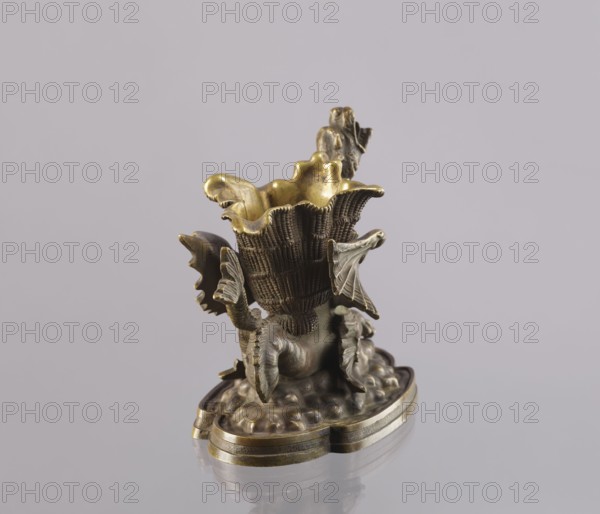 Spirit lamp, In the form of a two-winged dragon on a four-leaf oval base. Between the wings is a container; in the mouth is a hole for the wick, and on the side is a hole for pouring spirit. Described in a catalogue as an oil lamp and a very old one; however, because of the relatively large distance from the bottom of the container to the mouth poor capillarity for oil, it was most likely fired with spirit, and the container between the wings was used to hold cigarettes. 2nd half of the 19th century, Polesie. Lamp; bronze, — height 15.4 cm, width 20.8 cm, depth 10.5 cm., A small sculptural object sits on a reflective surface against a pale gray background, showing a textured cup-like form with scalloped edges and ornate leaf-like protrusions. The piece appears metallic with a mix of warm golden and darker bronze tones, with the interior and some raised edges showing brighter gold and other areas showing muted bronze. The base is rounded with an irregular, cloud-like platform and layere...