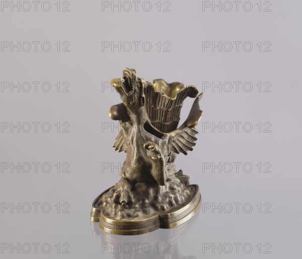 Spirit lamp, In the form of a two-winged dragon on a four-leaf oval base. Between the wings is a container; in the mouth is a hole for the wick, and on the side is a hole for pouring spirit. Described as an oil lamp but likely fired with spirit because of the distance from the container bottom to the mouth; the container between the wings was most likely used to hold cigarettes. 2nd half of the 19th century, Polesie. Lamp; height 15.4 cm, width 20.8 cm, depth 10.5 cm., A small metallic sculpture in bronze and brass tones sits on a reflective surface against a pale gray background; it depicts a stylized bird-like creature with an open beak, outstretched wings with feather details, clawed feet perched on a textured base, and a fluted vase-like element rising behind the creature, all showing varied bronze, brass, and dark brown patina.
