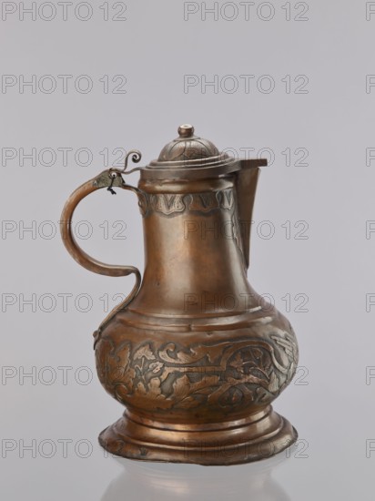 Jug. On a circular, stepped base, the body bulges in the lower part, decorated with a repoussé leafy twig and, at the rim, an eagle with spread wings. Above the two stepped areas is a neck with a spout. Repoussé leaves under the rim and along the rim. The hemispherical handle, fastened with rivets; the lid is hinged with a finger grip, arched, decorated with a leafy rosette and topped with a ball. Maker: Szewczykowski, Roman 1849–1901 attributed/role unknown. 1st half of the 18th century. Poland. Vessel. Height 39.5 cm; width 23.9 cm; depth 28.4 cm., A metal pitcher with a hinged domed lid and a curved handle sits against a plain pale gray background; the pitcher has a bulbous lower body, a tapered neck, a short spout, and a flared circular base, with decorative embossed floral and foliate patterns around the lower body and a band of ornamental motifs near the neck; the handle is attached with a small metal hinge and features a curled metal detail at the lid attachment; visible colors ...