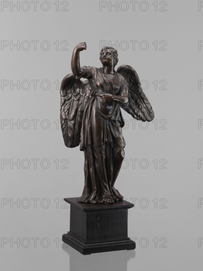 Statue - Angel, Figure of a standing angel with his right hand raised, the other raised to chest height, which originally contained a cross or an element of the Arma Christi. It is a pendant to another angel. Square wooden base with profiles on the upper and lower edges. Renaissance style, 16th century, sculpture; height 45 cm, width 12 cm, depth 12.5 cm., A dark brown statue of a winged figure stands on a black rectangular base against a pale gray background, showing sculpted drapery over the body, detailed feathered wings extending behind the shoulders, one arm raised and the other bent across the torso, with visible facial features, curly hair, and bare feet peeking from beneath flowing garments.