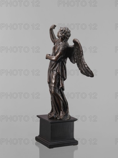 Statue - Angel, Renaissance, 16th century, Figure of a standing angel with his right hand raised, the other raised to chest height, which originally contained a cross or an element of the Arma Christi. It is a pendant to the latter. Square wooden base with profiles on the upper and lower edges, a secondary one. Sculpture, height 45 cm, width 12 cm, depth 12.5 cm., A dark brown metallic statue of a winged figure stands on a black square pedestal against a light gray background, showing a draped robe with flowing folds, curly hair, raised arm and an upward gaze, with textured feathered wings extending behind and bare feet visible at the base.