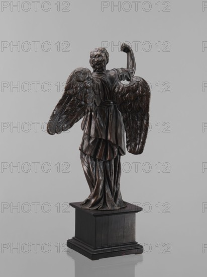 Statue - Angel, Renaissance, 16th century. Figure of a standing angel with his right hand raised, the other raised to chest height, which originally contained a cross or an element of the Arma Christi. It is a pendant to the latter. Square wooden base with profiles on the upper and lower edges, a secondary one. Sculpture, height 45 cm, width 12 cm, depth 12.5 cm., A dark brown wooden sculpture of a winged figure shown from the back standing on a dark rectangular pedestal, the figure wearing draped garments with carved folds and detailed feathered wings spread outward, the hair textured in curls and one arm raised with a closed hand, the surface showing polished highlights and deep shadows, set against a plain light gray background with reflections visible beneath the pedestal; the visible colors are dark brown, black, light gray, and subtle highlights of warm brown.