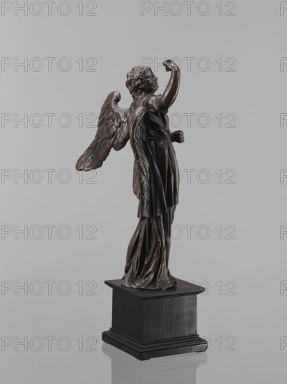 Statue - Angel. Figure of a standing angel with his right hand raised, the other raised to chest height, which originally contained a cross or an element of the Arma Christi. It is a pendant to the latter. Square wooden base with profiles on the upper and lower edges. Renaissance style, 16th century, sculpture, height 45 cm, width 12 cm, depth 12.5 cm., A dark bronze-colored statue of a winged figure stands on a black rectangular pedestal against a plain light gray background, the figure shown in profile with curly hair, draped garments, one arm raised and the other bent at the side, textured feathered wings extending from the back, and folds in the flowing clothing visible down to the base.