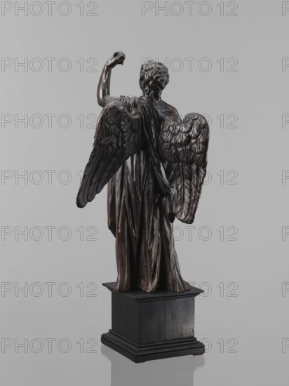 Statue — Angel, Renaissance; 16th century. Figure of a standing angel with his left hand raised, the other raised to chest height, which originally contained a cross or an Arma Christi element. Square wooden base with profiles on the upper and lower edges. Sculpture; height 44 cm, width 12 cm, depth 12.5 cm., A dark brown sculpture of a winged figure viewed from behind stands on a black rectangular plinth against a plain light gray background, the figure showing detailed textured wings and draped clothing with flowing folds, one arm raised and the other relaxed, hair rendered in sculpted curls, and the surface exhibiting varied brown and black tones with subtle highlights.