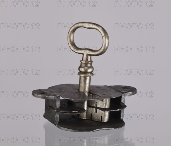 Register lock with key, locksmithing. Register on the bottom plate in the form of an irregular oval, with two semicircular projections with mounting holes that turn into flat supports for the cover in the form of a rectangle with rounded corners. Inside, a rod for the key guide made of double sheet metal; on both sides of it are single horizontal slats, and two on the bottom plate of the register. A rather primitive product. 19th century, Germany, lock; height 11.7 cm, width 16.3 cm, depth 5.3 cm., A close-up of a metal key inserted into a rectangular metal mechanism resting on a reflective surface, showing a rounded oval key head and a shaft with decorative ridges; the mechanism consists of layered, irregular metal plates and cutouts with visible seams and a few rough edges. The visible colors include silver, gray, dark gray, and muted beige, with subtle reflections and shadowed areas.
