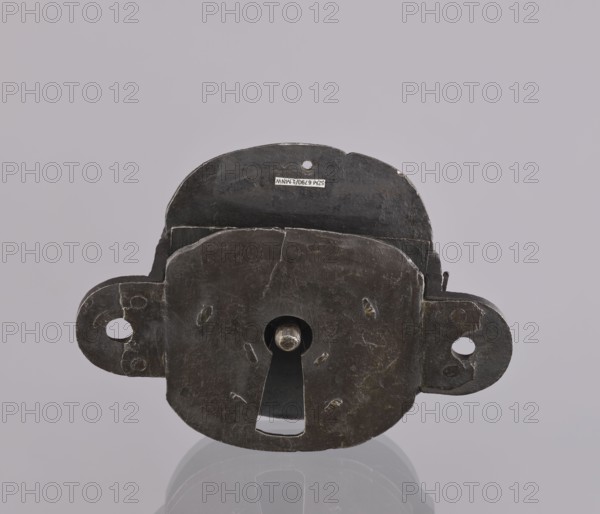 Lock register. The register on the bottom plate is in the form of an irregular oval, with two semicircular projections with mounting holes that turn into flat supports for the cover in the form of a rectangle with rounded corners. Inside the key rod is a guide made of double sheet metal; on both sides of it are single horizontal slats, and two on the bottom plate of the register. 19th century. Germany. Lock. Height 11.7 cm, width 16.3 cm, depth 5.3 cm., A dark metal lock plate with a rounded rectangular central body and two protruding rounded tabs on opposite sides, each tab featuring a circular hole, sits centered against a light gray background. The central body has a vertical keyhole with a circular upper opening and a tapered lower slot, and a short cylindrical metal pin visible through the upper opening. The metal surface shows irregular scratches, small dents, and worn edges with subtle lighter highlights and faint patches of brownish discoloration. The object casts a soft reflec...