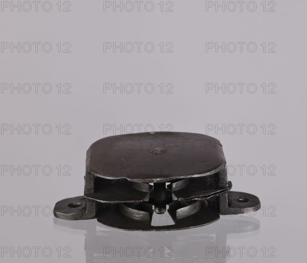 Lock register. The register on the bottom plate is in the form of an irregular oval, with two semicircular projections with mounting holes that turn into flat supports for the cover in the form of a rectangle with rounded corners. Inside the key rod is a guide made of double sheet metal; on both sides of it are single horizontal slats, and two on the bottom plate of the register. 19th century, Germany. Lock; height 11.7 cm, width 16.3 cm, depth 5.3 cm., A close-up of a small metallic object resting on a reflective surface, shown from a low frontal angle; the object is dark gray to black with a slightly worn, matte finish and rounded rectangular top edges, featuring layered metal plates and cutouts that reveal inner curved components and a central post; two flange-like extensions protrude sideways near the base, each with a round opening; the reflective surface beneath shows a faint mirrored outline of the object; the background is a smooth pale gray to light lavender.