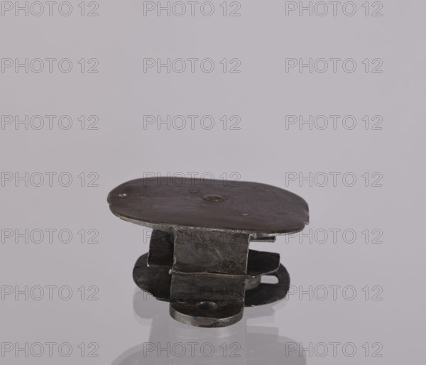 Lock register, 19th century, Germany. The register on the bottom plate is an irregular oval with two semicircular projections with mounting holes that turn into flat supports for the rectangular cover with rounded corners. Inside the key rod is a guide made of double sheet metal; on both sides are single horizontal slats, and two on the bottom plate of the register. Locksmithing; lock. Dimensions: height 11.7 cm, width 16.3 cm, depth 5.3 cm., A small, dark metallic object with a flat, rounded top and a layered base sits on a reflective surface; the metal appears worn and textured with a few small indents, and the visible colors are dark gray and black against a pale gray background with subtle reflections.