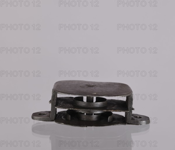Lock register. The register on the bottom plate is in the form of an irregular oval, with two semicircular projections with mounting holes that turn into flat supports for the cover in the form of a rectangle with rounded corners. Inside the key rod is a guide made of double sheet metal; on both sides of it are single horizontal slats, and two on the bottom plate of the register. 19th century, Germany. Lock; height 11.7 cm, width 16.3 cm, depth 5.3 cm., A small metal object sits centered on a reflective surface against a pale gray background; the object is dark gray to black with a flat, rounded top plate and a matching rounded base connected by short vertical supports, forming a layered, box-like shape with a visible circular opening and ring-like component in the middle, and a few mounting tabs extending outward on either side.
