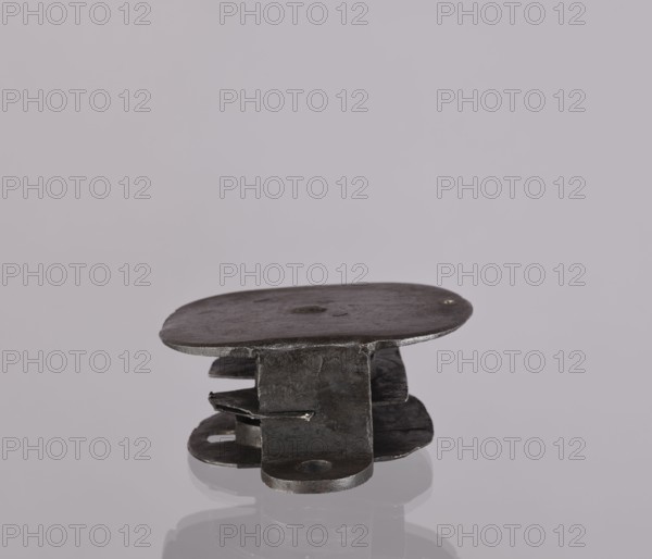 Lock register. The register on the bottom plate is in the form of an irregular oval, with two semicircular projections with mounting holes that turn into flat supports for the cover in the form of a rectangle with rounded corners. Inside the key rod is a guide made of double sheet metal; on both sides of it are single horizontal slats, and two on the bottom plate of the register. 19th century, Germany. Lock. Height 11.7 cm; width 16.3 cm; depth 5.3 cm. Maker/, A small metallic object sits on a reflective surface against a pale gray background; the object appears dark gray to black with a slightly uneven, textured surface and consists of layered, flat pieces stacked and fastened together, showing rounded and irregular edges and a few small punched holes; there are subtle highlights and shadowed areas on the metal, and the reflection below shows muted tones of gray and black.