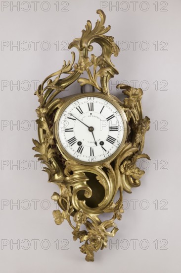 Cartel clock by Franciszek Gugenmus 1740–1820, 2nd half of the 18th century, Warsaw, Poland. Wall clock so-called cartel made of bronze with rocaille and floral motifs. White enameled dial with black Roman numerals, signed Fcois GUGENMUS / A VARSOVIE. Back covered with a brass sheet with rectangular doors. Mechanism unsigned; pendulum missing. Bronze casing without traces of gilding, possibly also made in Warsaw. Dimensions: height 80 cm, width 38 cm., A decorative wall clock with a white circular face set into an ornate gilded frame covered in carved scrolling leaves and floral motifs; the frame is predominantly gold with highlights where light reflects, and the clock face is white with dark hands and dark hour markers.