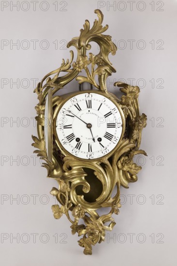 Cartel clock by Franciszek Gugenmus 1740–1820, 2nd half of the 18th century, Warsaw, Poland. Wall clock so-called cartel made of bronze with rocaille and floral motifs; bronze casing without traces of gilding, possibly made in Warsaw. White enameled dial with black Roman numerals, signed Fcois GUGENMUS / A VARSOVIE. Back covered with a brass sheet with rectangular doors. Mechanism unsigned; pendulum missing. Height 80 cm, width 38 cm., An ornate wall clock with an elaborate gilded frame featuring flowing rococo scrolls and floral motifs surrounds a round white clock face with black hands and black radial markers; the frame shows a gold color with highlights and shadowed areas, and the background is a pale light gray.