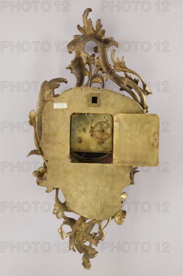 Cartel clock by Franciszek Gugenmus 1740–1820, 2nd half of the 18th century, Poland, Warsaw. Bronze wall clock so-called cartel with rocaille and floral motifs; white enameled dial with black Roman numerals, signed Fcois GUGENMUS / A VARSOVIE. Back covered with a brass sheet with rectangular doors; mechanism not signed and pendulum missing. Bronze casing without traces of gilding, possibly made in Warsaw. Height 80 cm, width 38 cm., An ornate metallic object with a decorative leafy frame and scrollwork in gold tones surrounds a central flat plate with a square opening and a hinged door ajar, revealing inner mechanical components with a patina of greenish and darker metallic hues; the frame and plate show areas of worn and varied gold, with some darker tarnished spots, and the background is a pale neutral color.