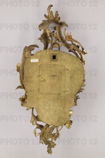Cartel clock by Franciszek Gugenmus 1740–1820, 2nd half of the 18th century, Poland Warsaw. Bronze wall clock so-called cartel with rocaille and floral motifs; bronze casing without traces of gilding, possibly also made in Warsaw. White enameled dial with black Roman numerals, signed Fcois GUGENMUS / A VARSOVIE. Back covered with a brass sheet with rectangular doors. Mechanism not signed; pendulum missing. Height 80 cm, width 38 cm., A decorative metallic object photographed against a neutral light background, showing an ornate back with flowing, scroll-like floral and leaf forms and a smooth central panel with a small recessed square opening; the object is predominantly gold in color with areas of darker tarnish and subtle brownish patina, and the background is pale gray.