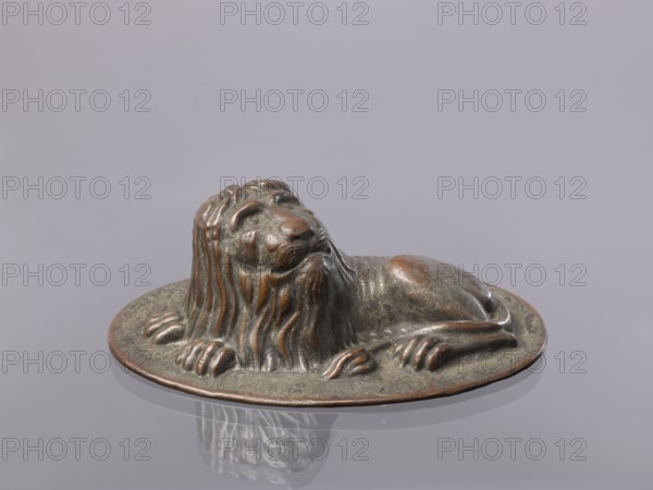 Jelly mold, repoussé copper sheet in the form of a lying lion; raised head allows contents to be removed intact., described variously as a button or jelly mold. 18th–19th century, possibly Germany. Vessel; height 5.4 cm, width 15.5 cm, depth 9.8 cm., A small sculpted lion figure resting on an oval base, showing a reclining pose with its head raised and mane flowing down its neck and chest; the surface of the sculpture appears metallic with a mix of greenish patina and brownish-bronze highlights, and the base shares the same coloration; the background is a smooth, light grayish-lavender surface that reflects the sculpture faintly.