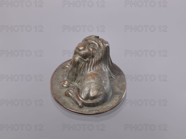 Jelly mold, repoussé copper sheet in the form of a lying lion; raised head allows contents to be removed intact. Possibly 18th–19th century, Germany, vessel; height 5.4 cm, width 15.5 cm, depth 9.8 cm., A small sculpted object showing a reclining lion with a flowing mane positioned on a shallow circular base, rendered in metallic tones of brown, dark bronze, and subtle greenish patina, sitting on a smooth pale gray surface; the lion's form shows rounded limbs, textured mane ridges, and a slightly raised head with closed features, and the base has irregular discoloration and reflective highlights.