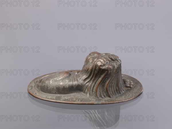 Jelly mold, repoussé copper sheet in the form of a lying lion with raised head allowing contents to be removed intact. Probably 18th–19th century, Germany, vessel; height 5.4 cm, width 15.5 cm, depth 9.8 cm., A small sculpted object resembling a reclining animal with flowing mane-like ridges sits on an oval base; the surface shows a weathered metallic finish with greenish-gray and brownish-copper tones, with patches of darker and lighter areas and a subtle reflective sheen on the base, all set against a smooth pale gray background.