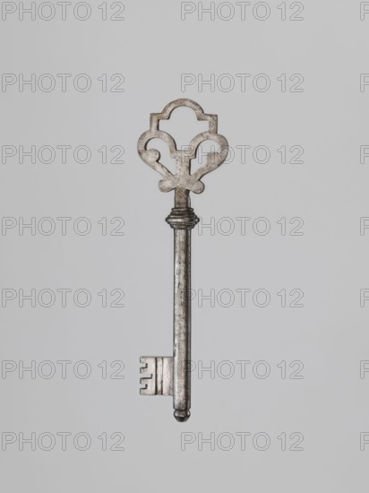 Key, Roman Szewczykowski 1849–1901 provenance gift; 19th century locksmithing key. Full shaft ending in a flattened ball, with a quill with cutouts on the lower edge for a ratchet lock. Base of the shaft with a profiled bulge; handle in the form of a heart topped with a p-rectangle with a hemispherical upper edge. Height 14.1 cm; width 4 cm; depth 1.5 cm., A single ornate metal key is centered against a light gray background; the key has a decorative bow with curved cutouts and a weathered silver-gray surface, a long cylindrical shank with subtle darker patina, a ridged collar near the top, and a rectangular bit with several notches, ending in a rounded tip.
