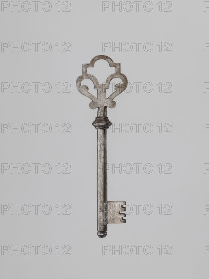 Key, Roman Szewczykowski 1849–1901 — 19th century. Locksmithing key with a full shaft ending in a flattened ball, featuring a quill with cutouts on the lower edge for a ratchet lock. Base of the shaft has a profiled bulge; handle is heart-shaped topped with a p-rectangle with a hemispherical upper edge. Dimensions: height 14.1 cm, width 4 cm, depth 1.5 cm., A single ornate metal key is centered against a plain light gray background, showing a decorative heart-shaped bow with symmetrical cutouts and a slightly worn, mottled silver-gray surface, a long cylindrical shaft with a rounded tip and a small decorative collar near the top, and a rectangular bit with three notches; overall colors visible include shades of silver-gray and light gray.